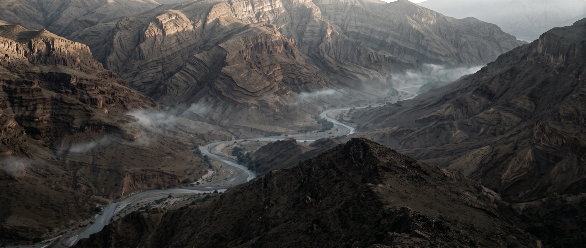A wadi in the Hajar mountains seen from a high ridge at dawn, layered rock and a winding riverbed